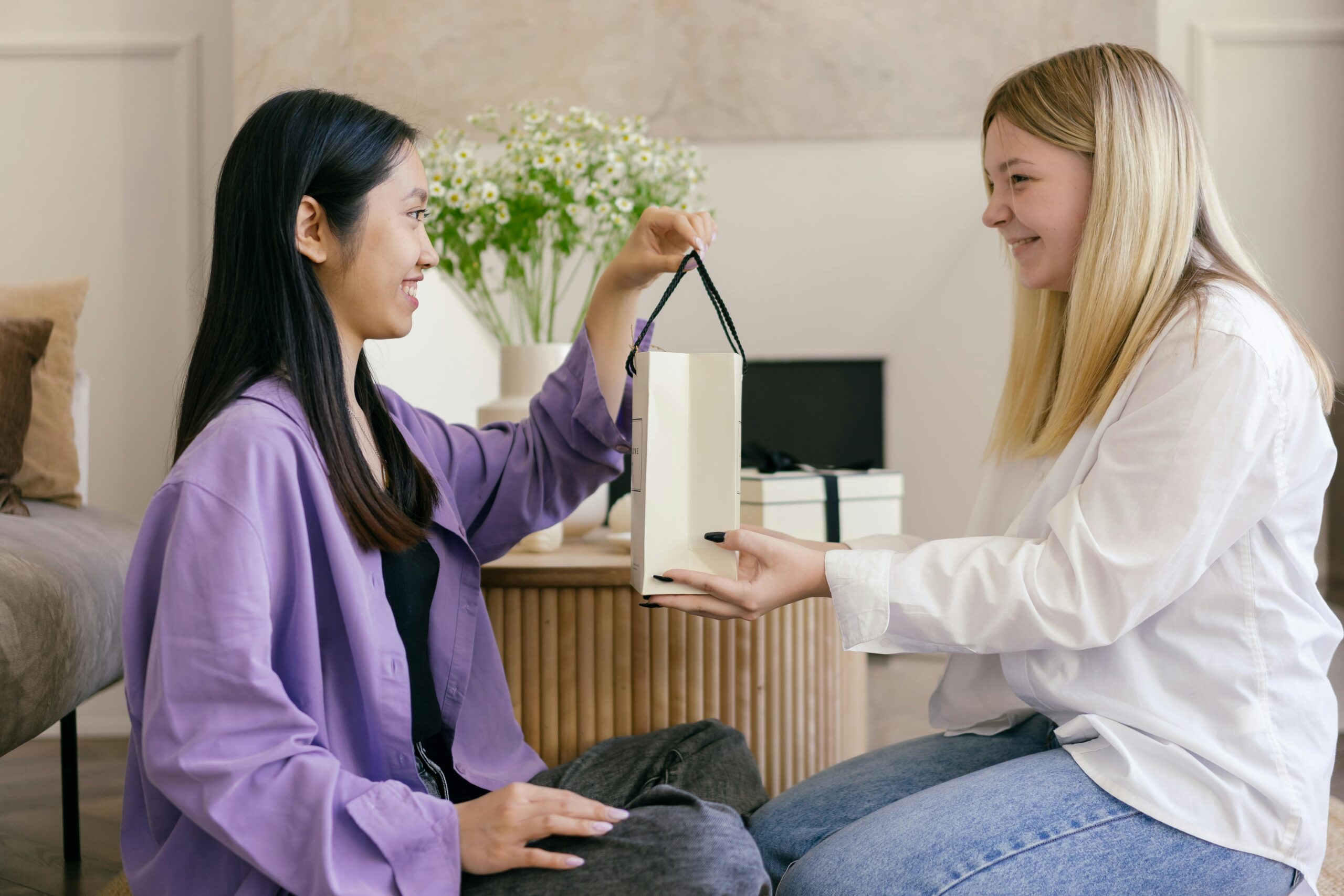 Two women smiling and exchanging a gift indoors, symbolizing friendship and celebration.